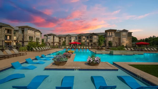 Resort-style pool with built-in lounge areas surrounded by an expansive sundeck with poolside lounging, private cabanas, and the apartment homes and a beautiful sunset in the background